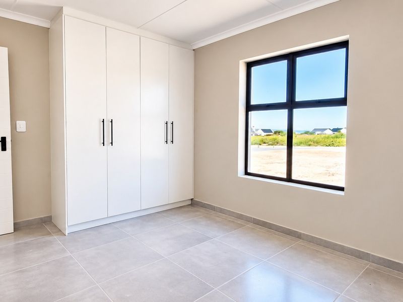 A bright, modern bedroom featuring neutral walls, large-format floor tiles, and ample built-in cupboard storage with a view through the window.