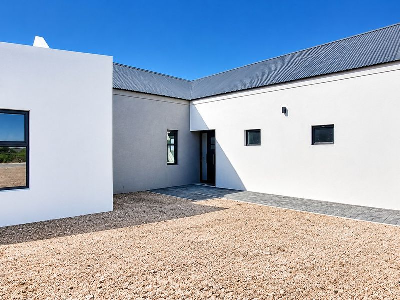 Modern single-story home exterior featuring clean white and grey rendered walls, corrugated metal roofing, and a neat gravel and paved entrance area.
