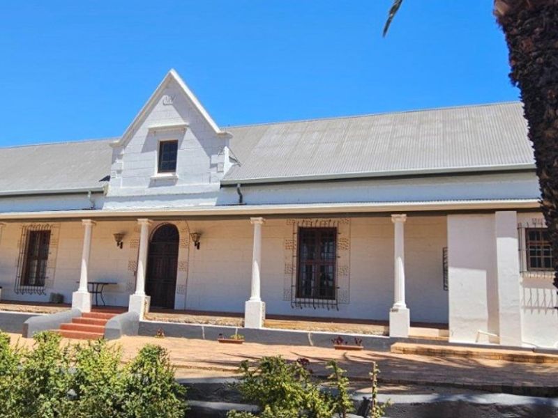 A well-maintained farmhouse featuring a classic white facade, a covered veranda with columns, and a corrugated iron roof, set in a rural landscape.