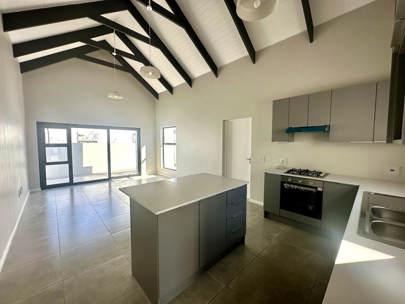 Modern open-plan kitchen and living area featuring a vaulted ceiling with exposed beams, sleek cabinetry, a central island, and direct access to the patio.
