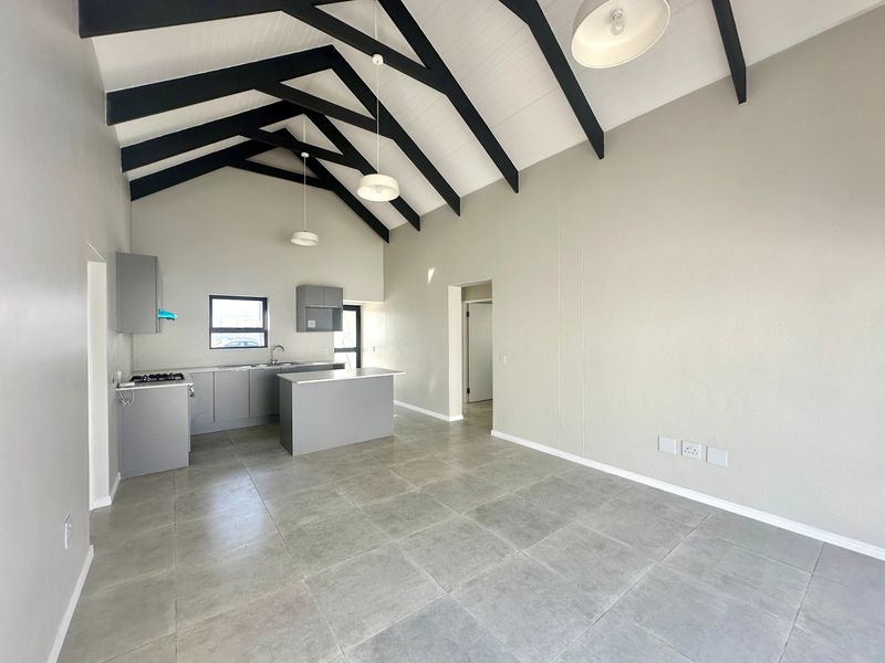 Spacious open-plan living area featuring a high vaulted ceiling with exposed beams, modern grey kitchen cabinetry, and neutral tiled flooring throughout.