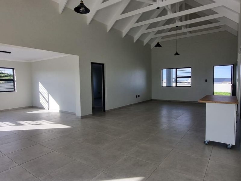 Spacious open-plan living area featuring high vaulted ceilings with exposed rafters, neutral tiled flooring, and abundant natural light through large windows.