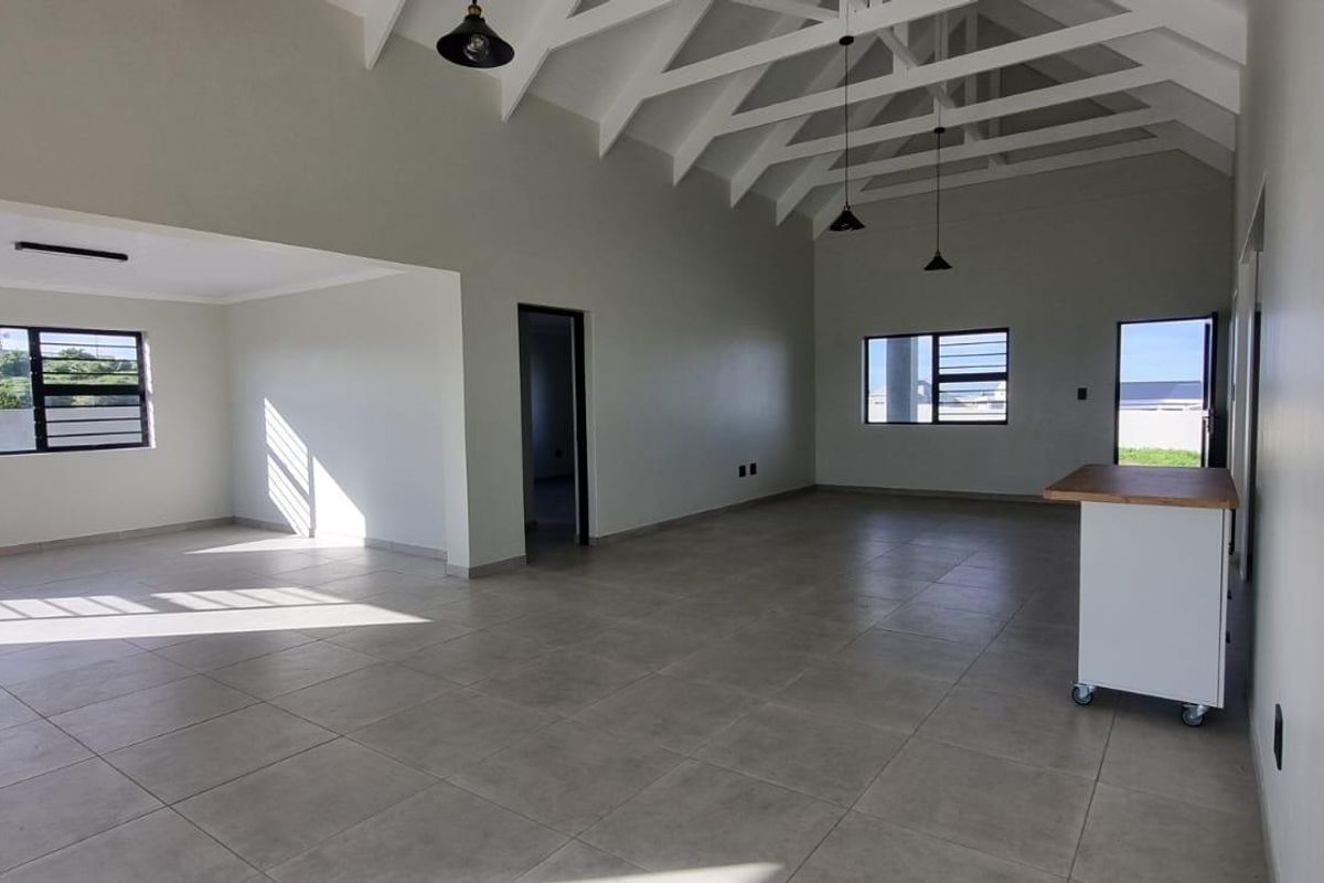 Spacious open-plan living area featuring high vaulted ceilings with exposed rafters, neutral tiled flooring, and abundant natural light through large windows.