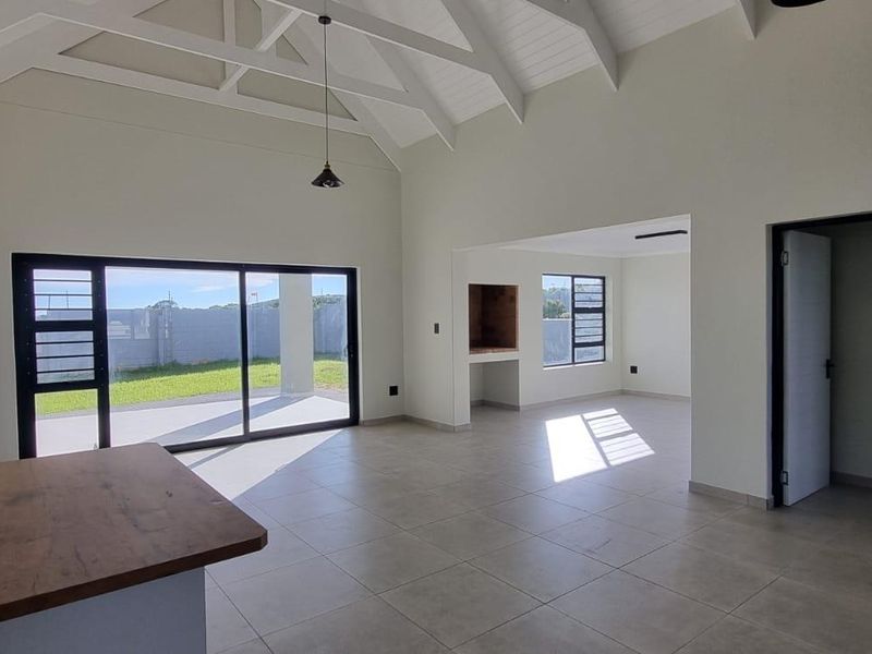 Spacious open-plan living area featuring a vaulted ceiling with exposed trusses, tiled floors, and a built-in braai, leading out to a patio.