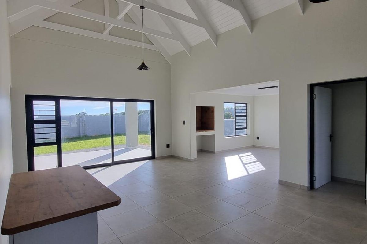 Spacious open-plan living area featuring a vaulted ceiling with exposed trusses, tiled floors, and a built-in braai, leading out to a patio.