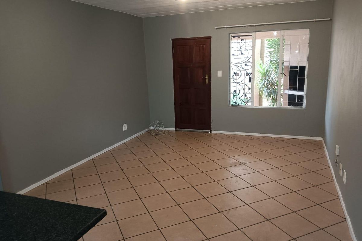 An empty, open-plan living area featuring neutral grey walls, tiled flooring, and a secure timber entrance door with window security bars.