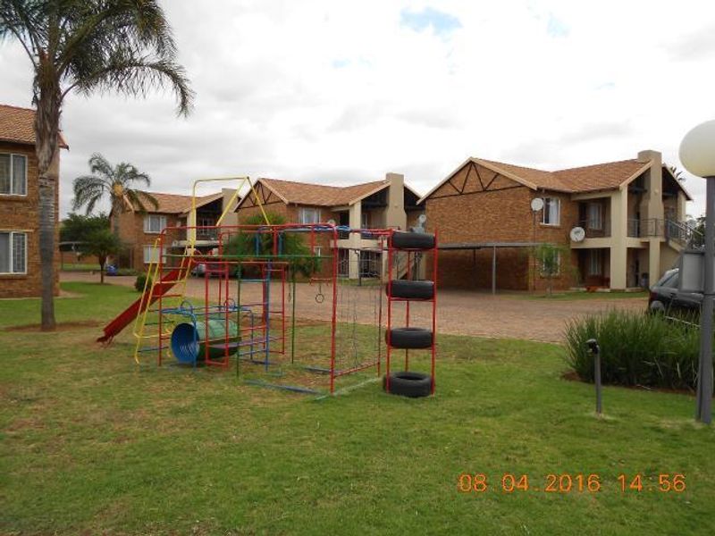 Exterior view of a townhouse complex featuring brick facades, tiled roofs, a paved driveway, and a communal garden area with playground equipment.