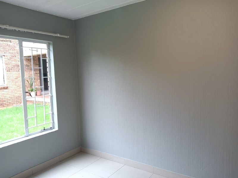 A clean, empty bedroom featuring neutral grey walls, tiled flooring, and a window fitted with security bars overlooking the garden.