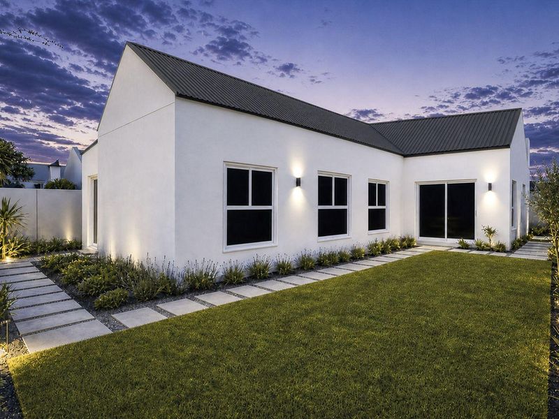 A beautifully maintained rear exterior view of a modern home featuring a clean white facade, dark metal roof, and landscaped garden with paved pathways.