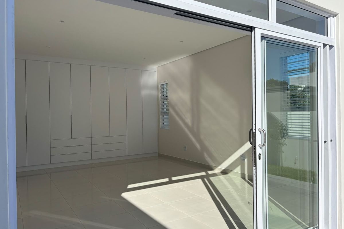 A bright, empty bedroom featuring modern white built-in cupboards, large format floor tiles, and a wide sliding glass door opening to the exterior.
