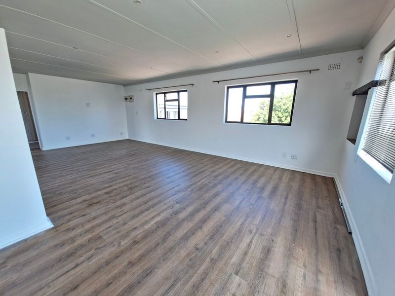 A spacious, empty living area featuring modern laminate flooring, neutral white walls, and large windows that provide ample natural light.
