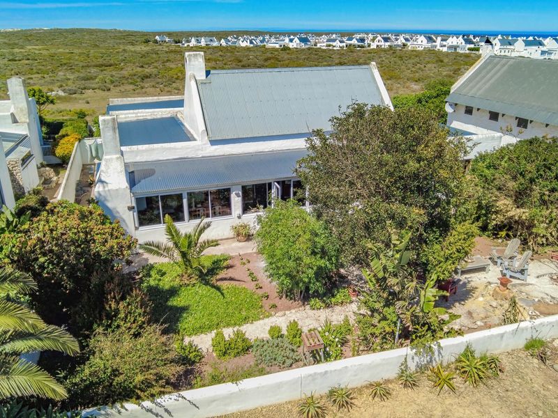 Aerial view of a coastal property featuring a white house with a corrugated metal roof, large glass doors, and a mature landscaped garden.
