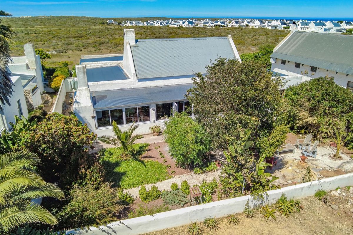 Aerial view of a coastal property featuring a white house with a corrugated metal roof, large glass doors, and a mature landscaped garden.