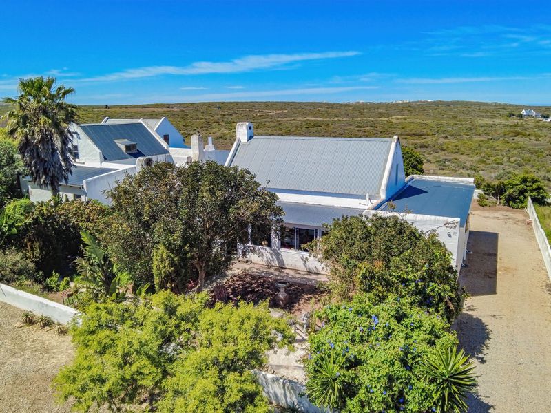 Aerial view of a coastal property featuring a white house with a corrugated metal roof, surrounded by established trees and a gravel driveway.