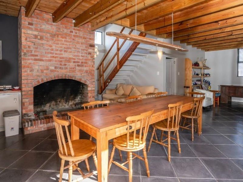 Open plan living and dining area featuring exposed timber ceiling beams, a prominent brick fireplace, and a staircase with a timber handrail.