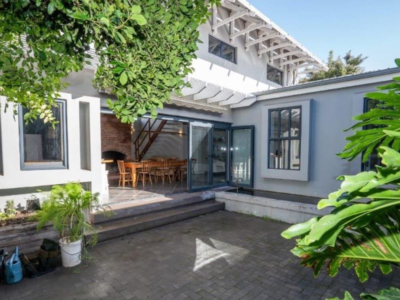 Rear exterior view featuring a paved courtyard, timber pergola, and seamless indoor-outdoor flow through bi-fold doors into a living space with a brick fireplace.