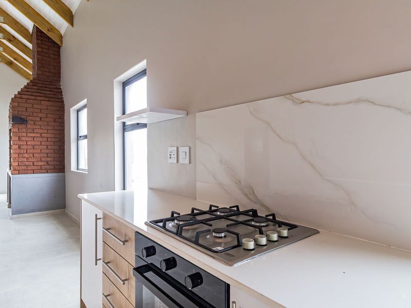 Modern kitchen area featuring a gas hob, built-in oven, marble-look splashback, and high ceilings with exposed timber trusses and a feature brick fireplace.