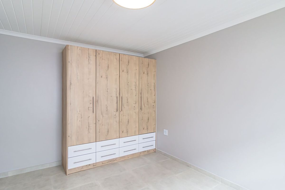 A clean, neutral bedroom featuring light-toned tiled flooring, a tongue and groove ceiling, and a large, modern built-in wardrobe with integrated drawer storage.