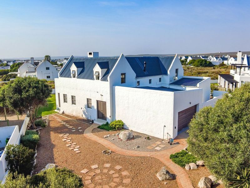 Aerial view of a white coastal-style house with a metal roof, featuring a landscaped garden, paved pathways, and an attached garage.