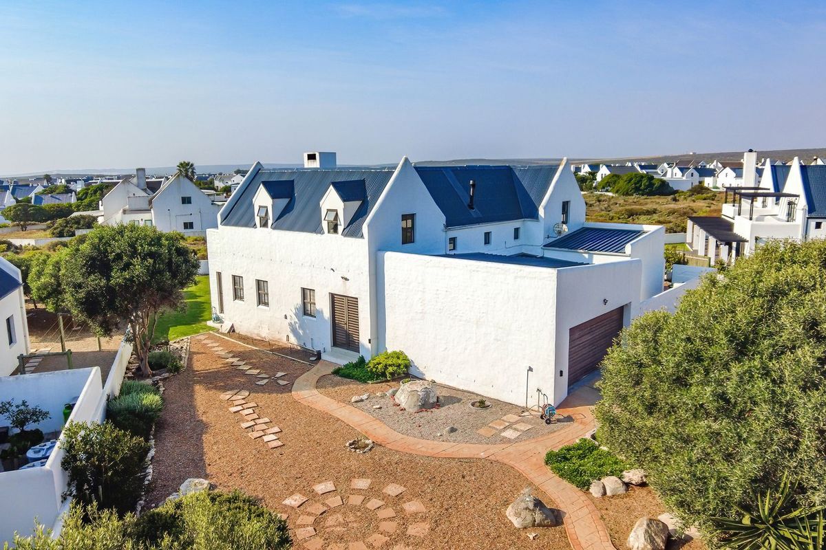 Aerial view of a white coastal-style house with a metal roof, featuring a landscaped garden, paved pathways, and an attached garage.
