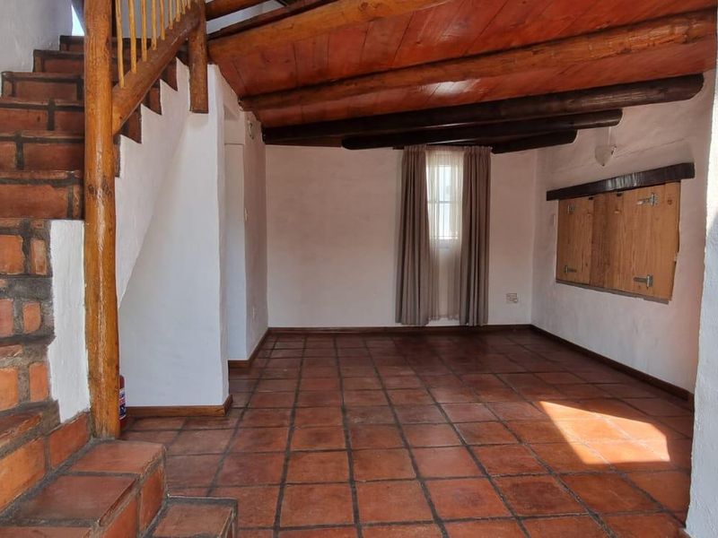 An empty living area featuring rustic terracotta tile flooring, exposed timber ceiling beams, and a wooden staircase with a matching balustrade.