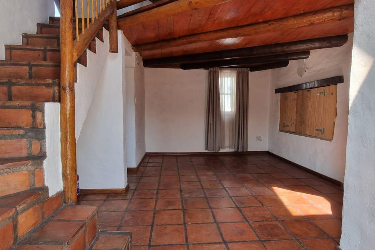 An empty living area featuring rustic terracotta tile flooring, exposed timber ceiling beams, and a wooden staircase with a matching balustrade.