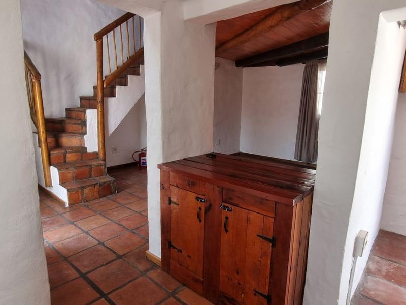 A rustic hallway featuring terracotta floor tiles, a wooden staircase with a matching handrail, and exposed timber ceiling beams.