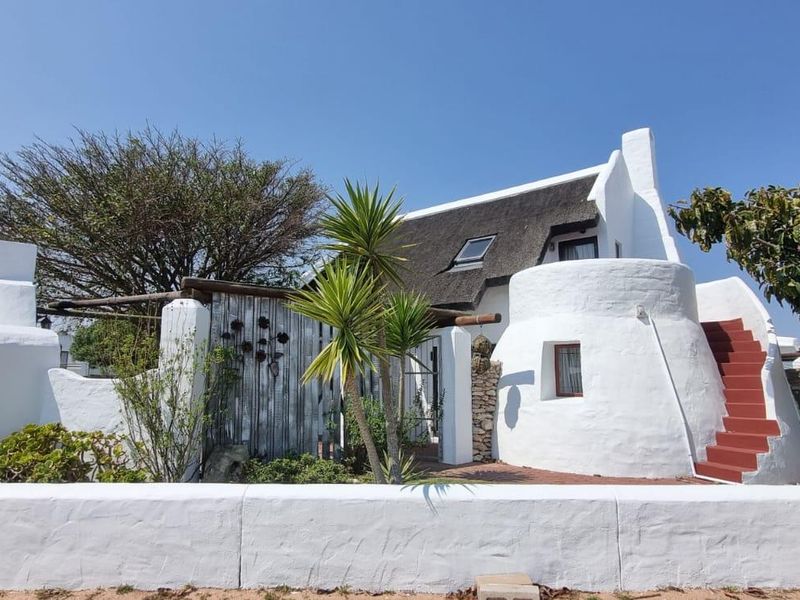 A charming white-washed cottage featuring a traditional thatched roof, a distinctive curved facade with an external staircase, and a private enclosed garden area.