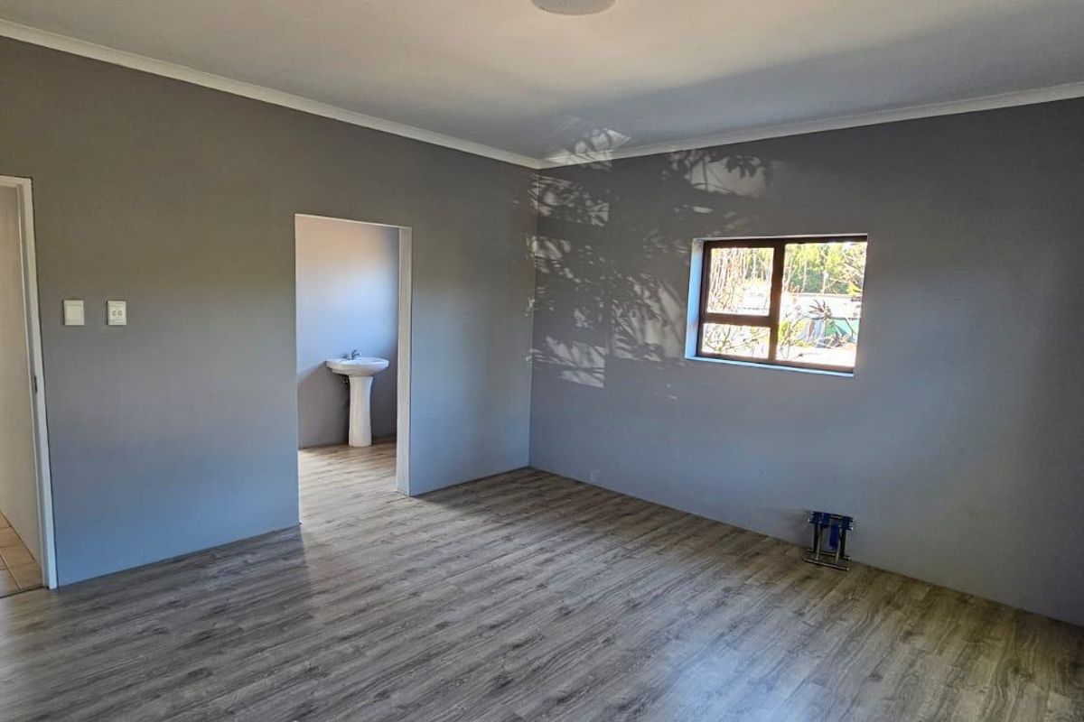 A spacious, empty bedroom featuring modern laminate flooring, neutral grey walls, and an adjoining en-suite area with a pedestal basin.