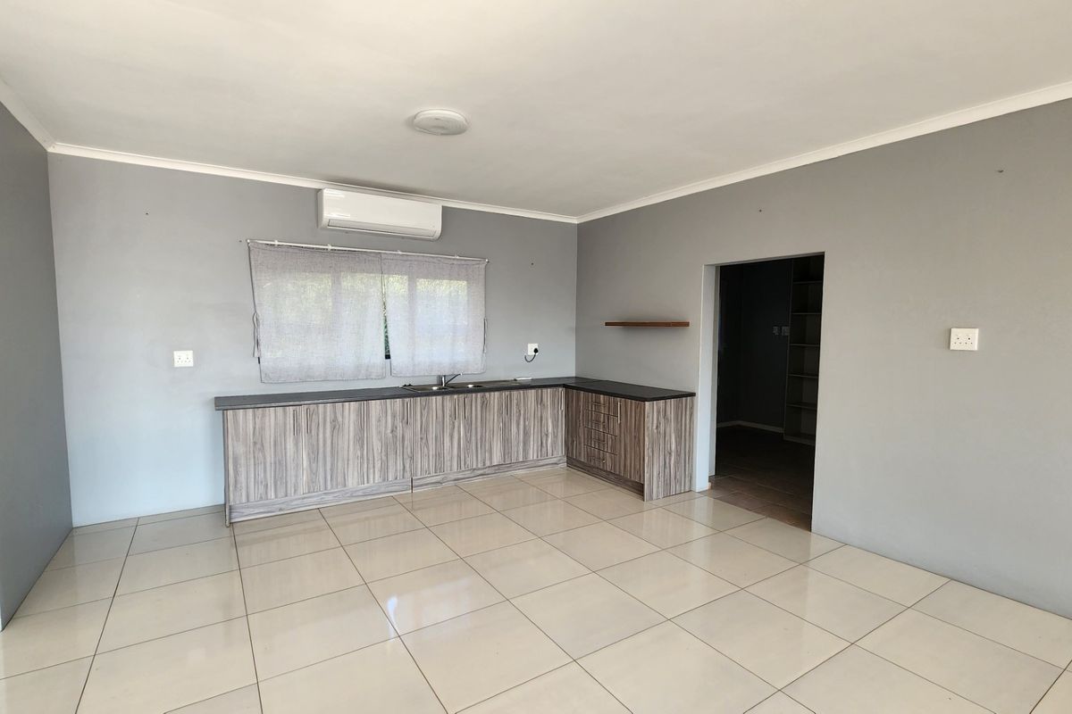 A clean, open-plan kitchen area featuring modern wood-look cabinetry, dark countertops, tiled flooring, and a wall-mounted air conditioning unit.