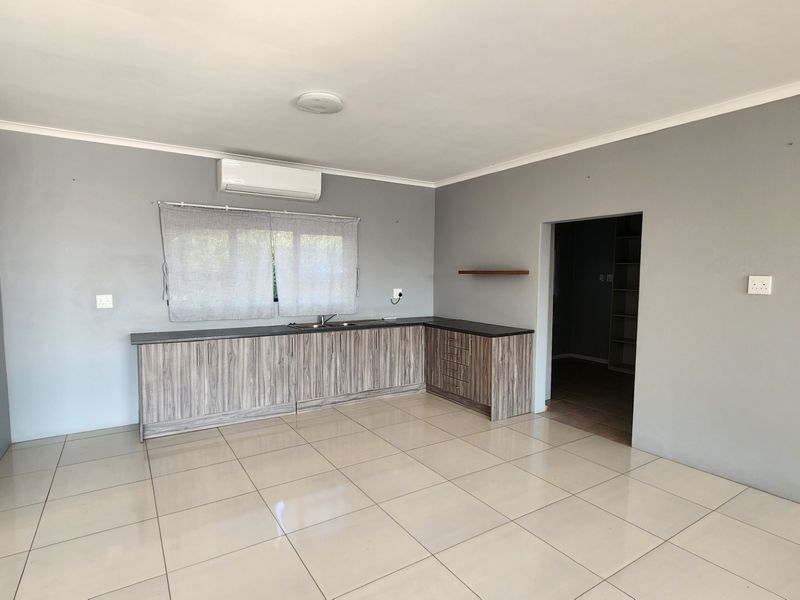 A clean, open-plan kitchen area featuring modern wood-look cabinetry, dark countertops, tiled flooring, and a wall-mounted air conditioning unit.