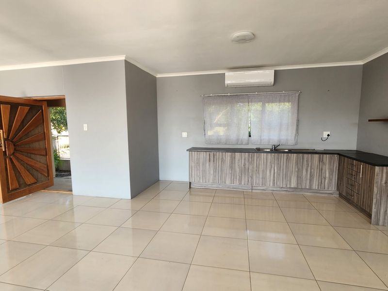 An open-plan living and kitchen area featuring neutral grey walls, tiled flooring, modern wood-grain cabinetry, and a wall-mounted air conditioning unit.