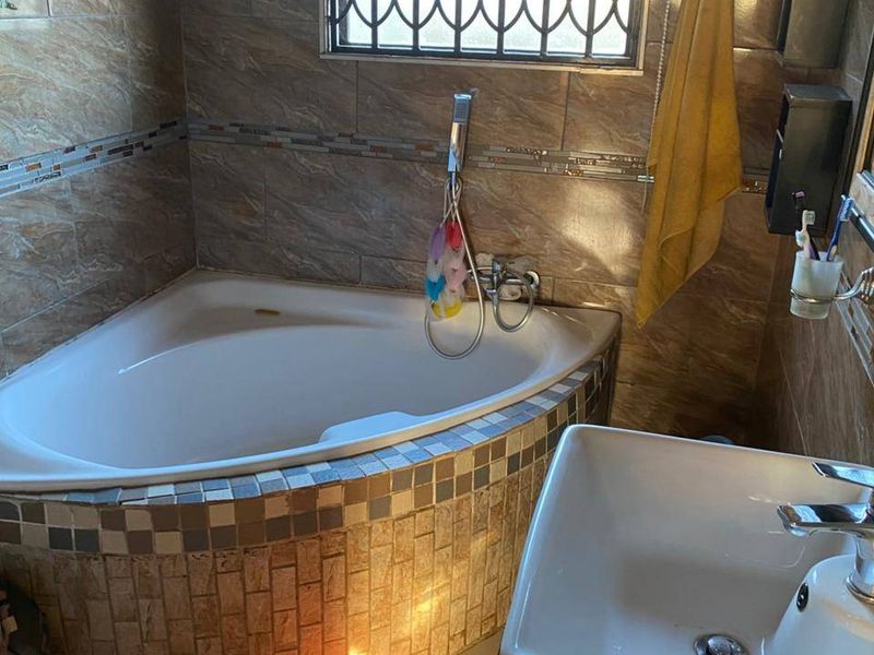 A bathroom featuring a corner bathtub with tiled surround, a modern basin, and a window fitted with security burglar bars.