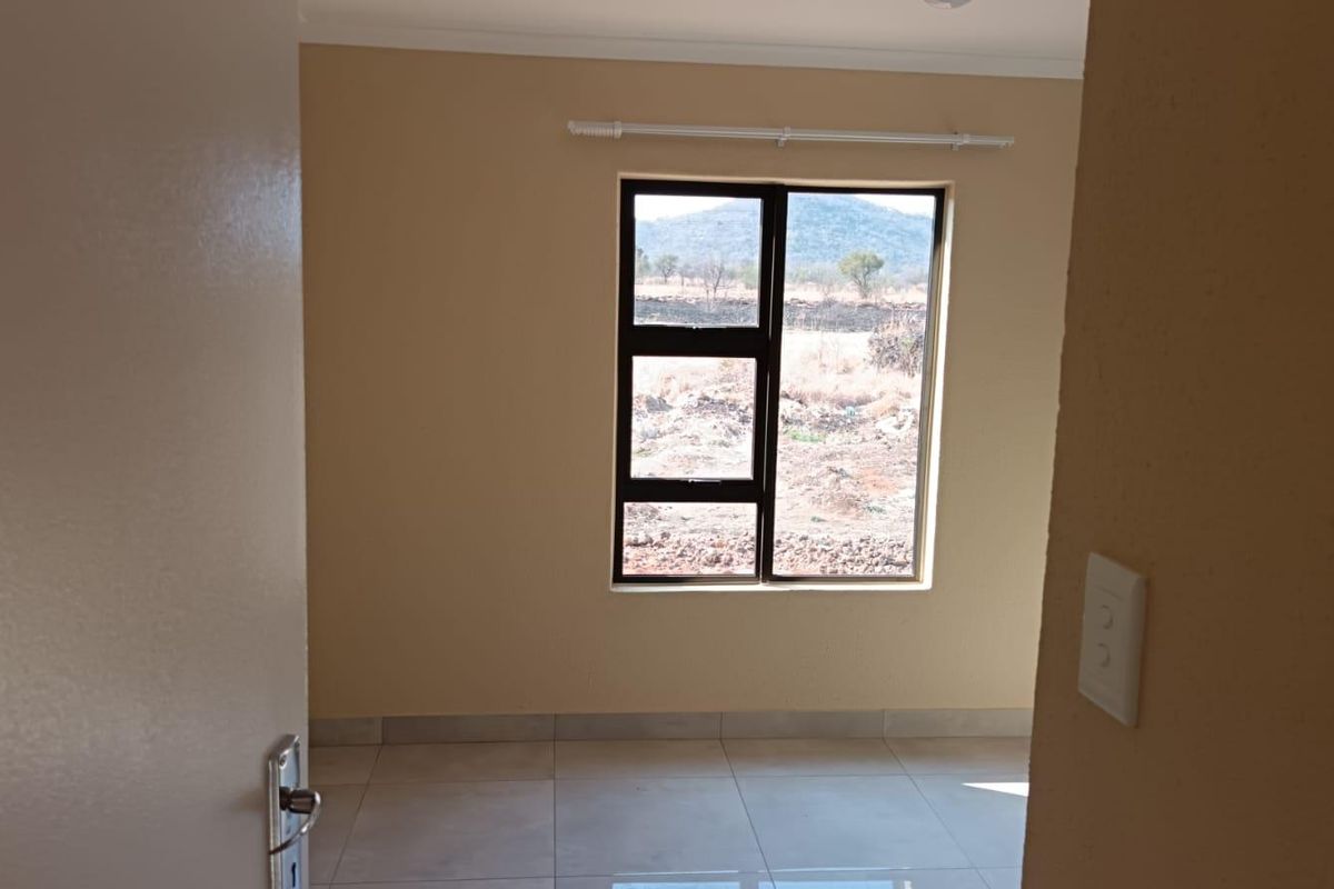 A bright, empty bedroom featuring neutral wall paint, tiled flooring, and an aluminium window frame with views of the surrounding landscape.