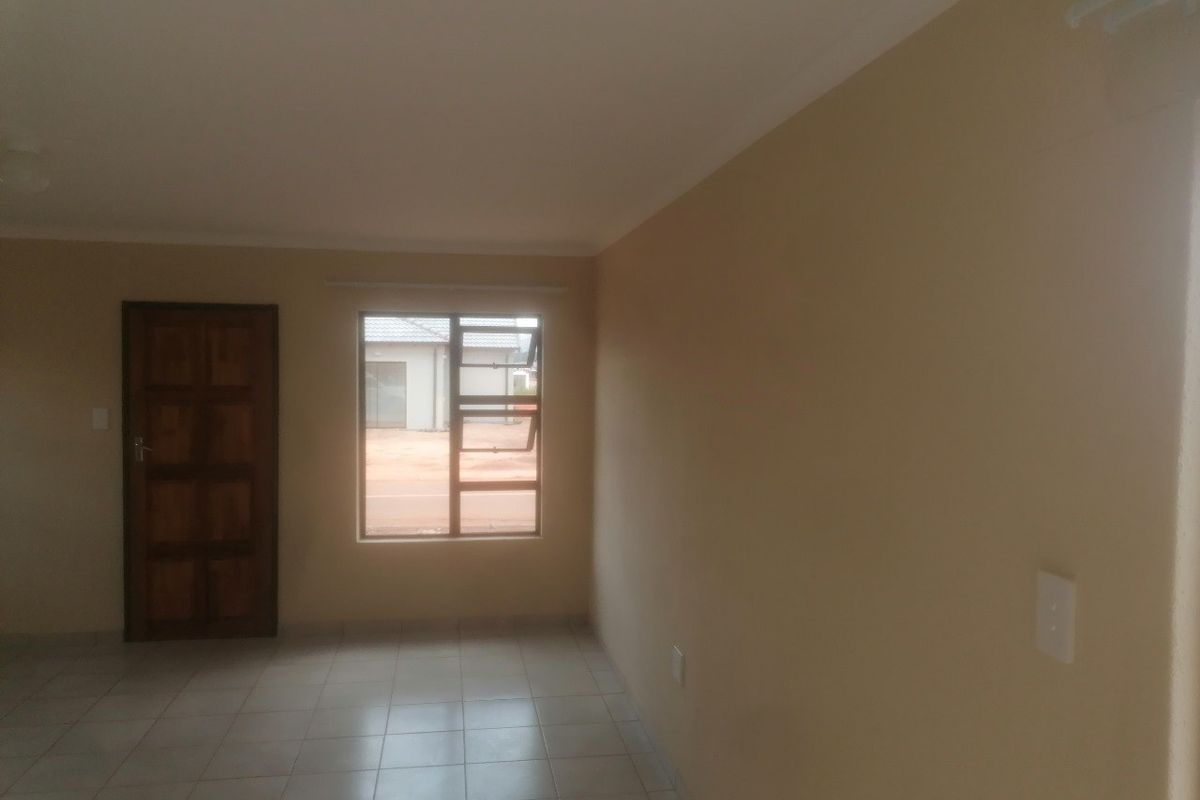 A bright, empty living room featuring neutral wall tones, tiled flooring, and a timber entrance door with a view to the exterior.