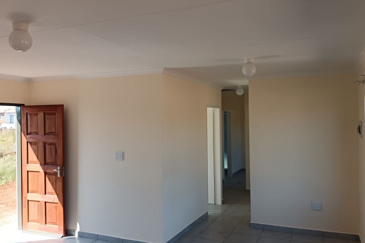 A bright, empty living area featuring neutral walls, tiled flooring, and direct access to the exterior, leading into a central hallway.