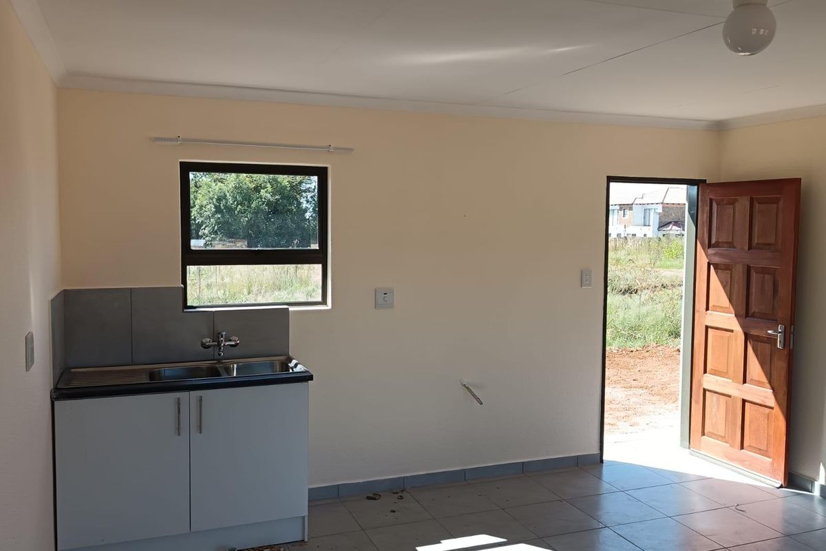 A compact, empty kitchen area featuring a sink unit, tiled flooring, and direct access to the exterior through a timber door.