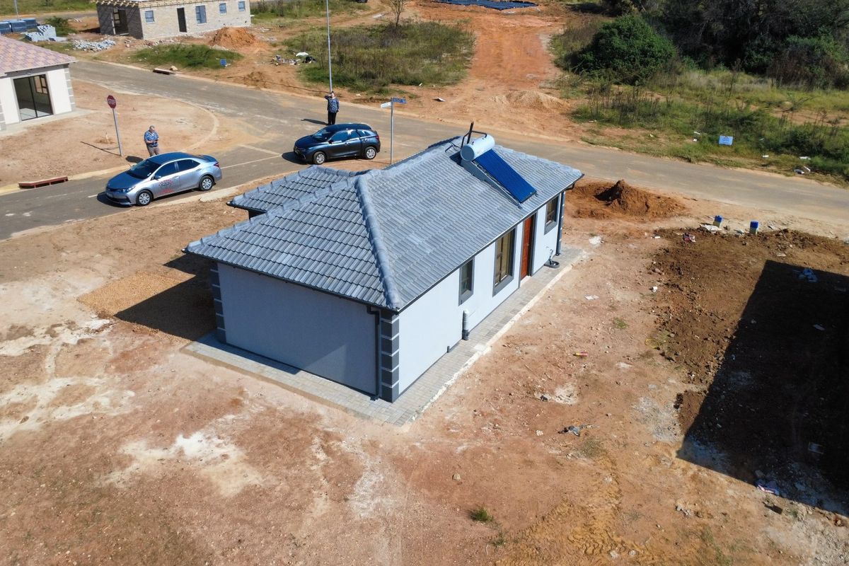 Aerial view of a newly constructed single-story house featuring a tiled roof, solar water heater, and paved perimeter on an unlandscaped plot.
