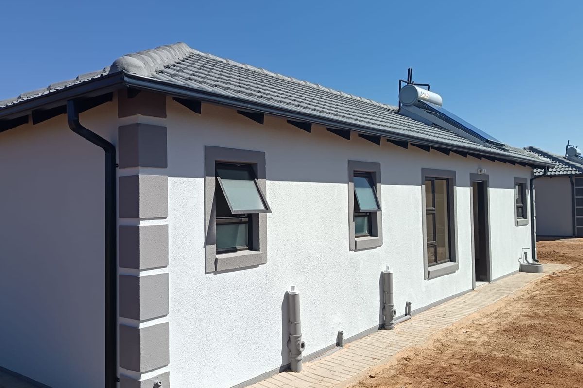 Rear exterior view of a modern house featuring a tiled roof, solar water heating system, and paved walkway along the building perimeter.