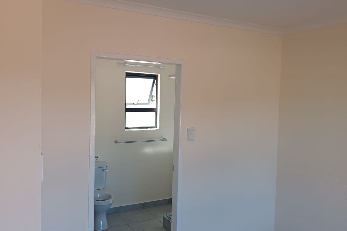 A view from an adjacent room into a simple, functional bathroom featuring tiled flooring, a toilet, and a window.