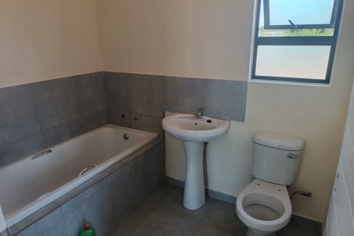 A functional bathroom featuring a bathtub, pedestal basin, and toilet with grey tiled finishes and a top-hung window.