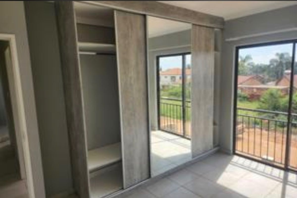 A bright, empty bedroom featuring modern built-in wardrobes with sliding mirror doors, tiled flooring, and direct access to a balcony.