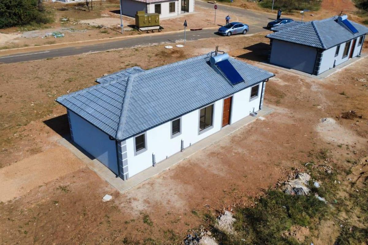 Aerial view of a newly constructed single-story house featuring a tiled roof with an integrated solar water heating system and surrounding unlandscaped grounds.