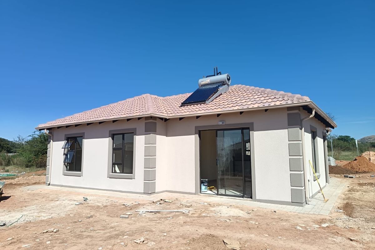 Rear view of a modern single-story house featuring a tiled roof, solar water heating system, and sliding glass doors, set on an unfinished plot.