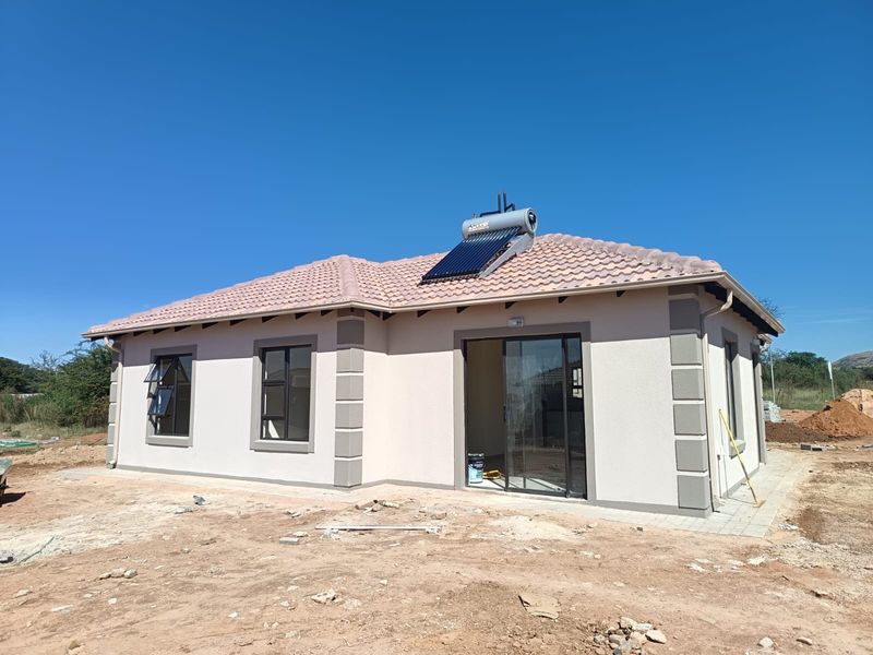Rear view of a modern single-story house featuring a tiled roof, solar water heating system, and sliding glass doors, set on an unfinished plot.