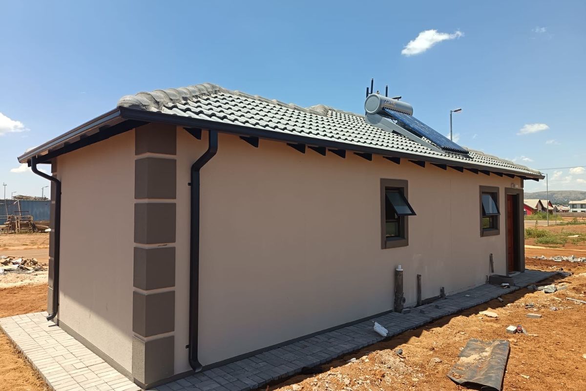 Rear view of a modern house featuring a solar water heater, tiled roof, and paved perimeter walkway in a developing residential area.