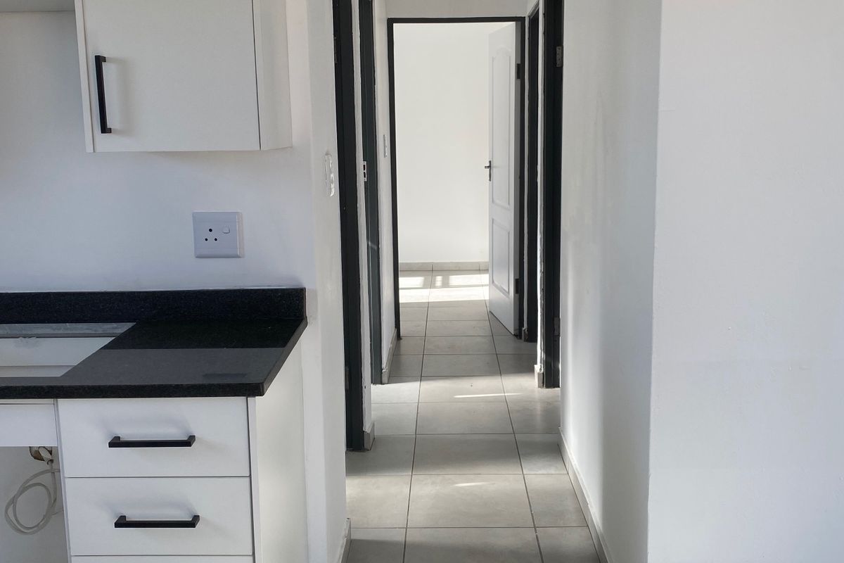 A clean, modern kitchen area featuring white cabinetry, dark stone countertops, and tiled flooring, leading into a bright hallway.