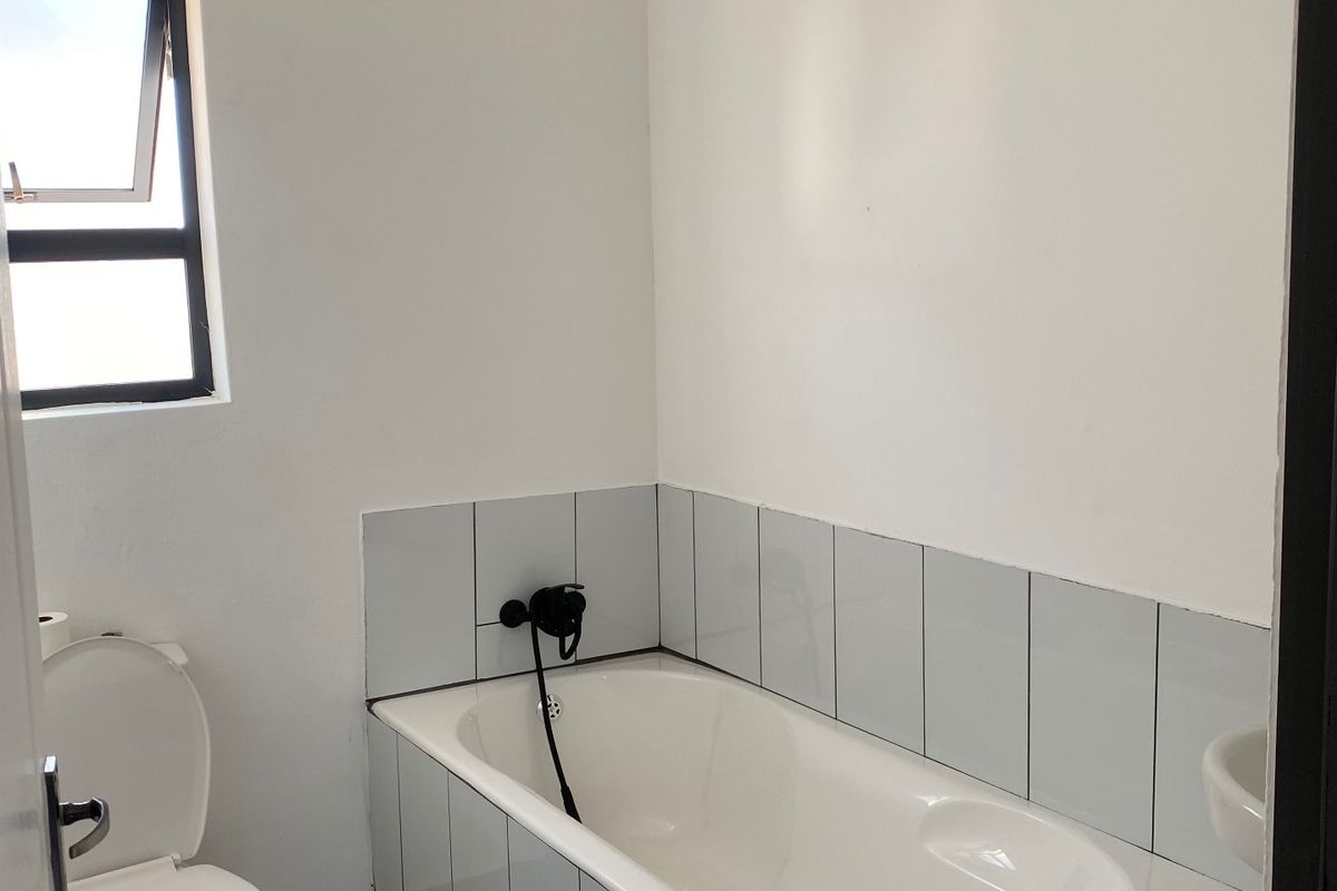 A functional bathroom featuring a bathtub with black tapware, a toilet, and neutral tiled finishes, offering a clean and simple layout.
