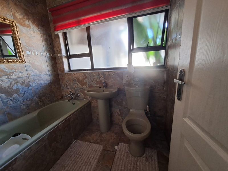 A functional bathroom featuring a pedestal basin, toilet, and built-in bathtub with full-height wall tiling and natural light from an aluminium-framed window.