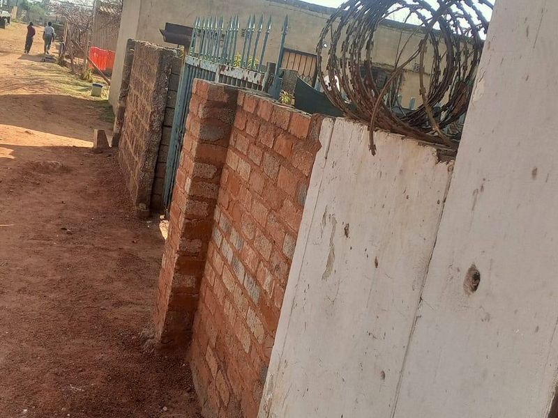 A view of the property boundary featuring a brick wall, metal gate, and security razor wire along an unpaved street.